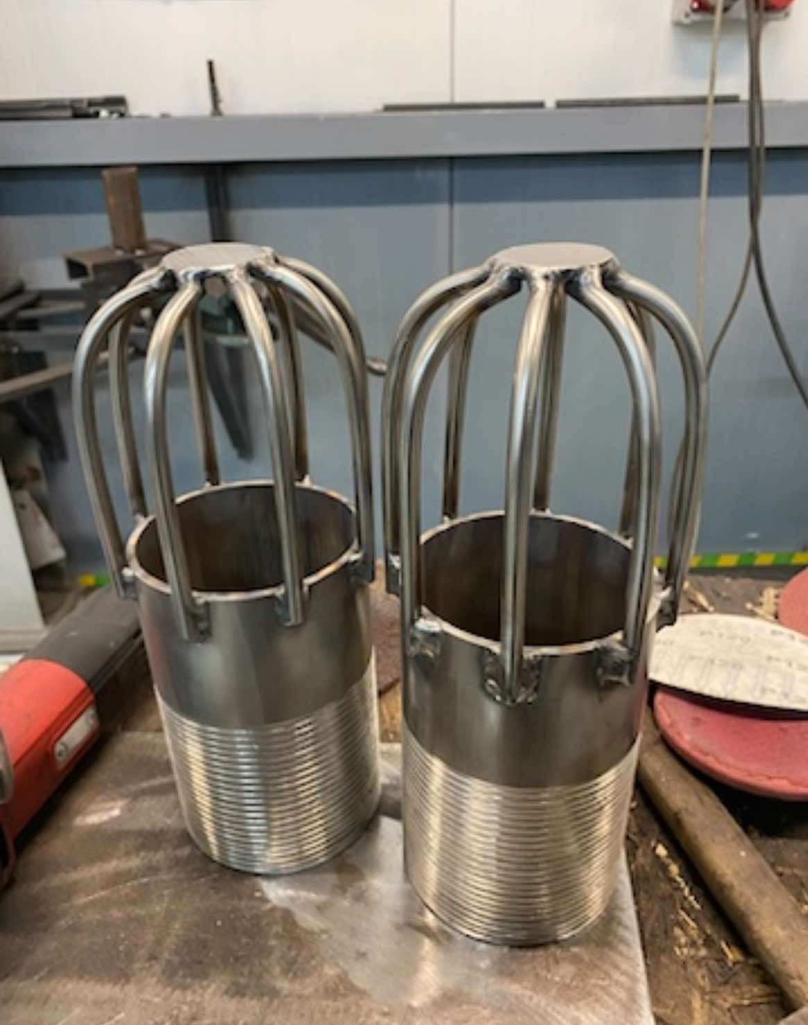 Dwa metalowe cylindry z prętami tworzącymi klatkę, leżące na warsztatowym stole. Widoczne ślady obróbki i spawania. W tle narzędzia i elementy konstrukcyjne.