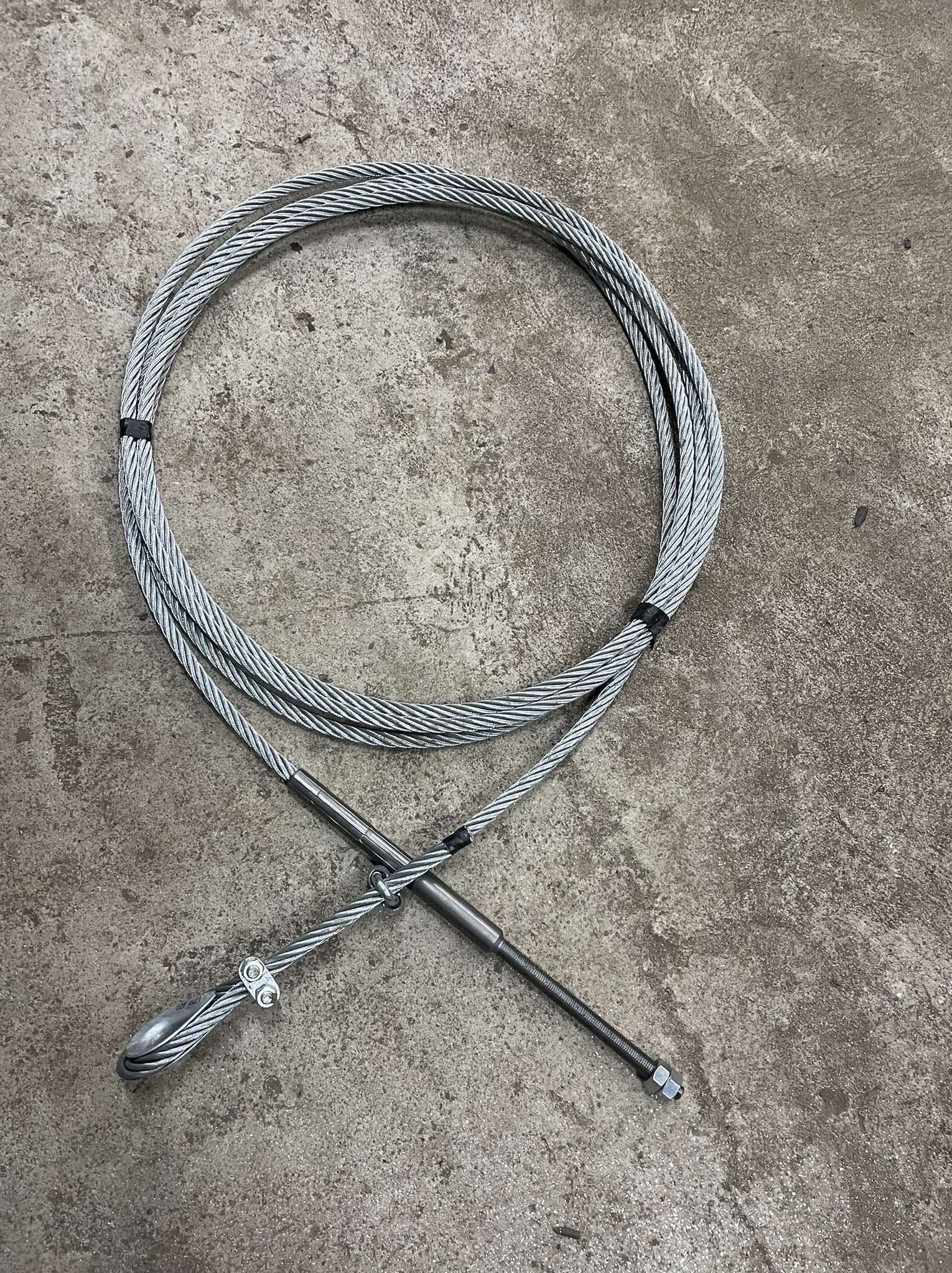 Stalowa lina z zaciskami i gwintowanym prętem na szarym, betonowym tle. Lina zwinięta w okrąg, zabezpieczona czarną taśmą. Widoczne detale mocowań.