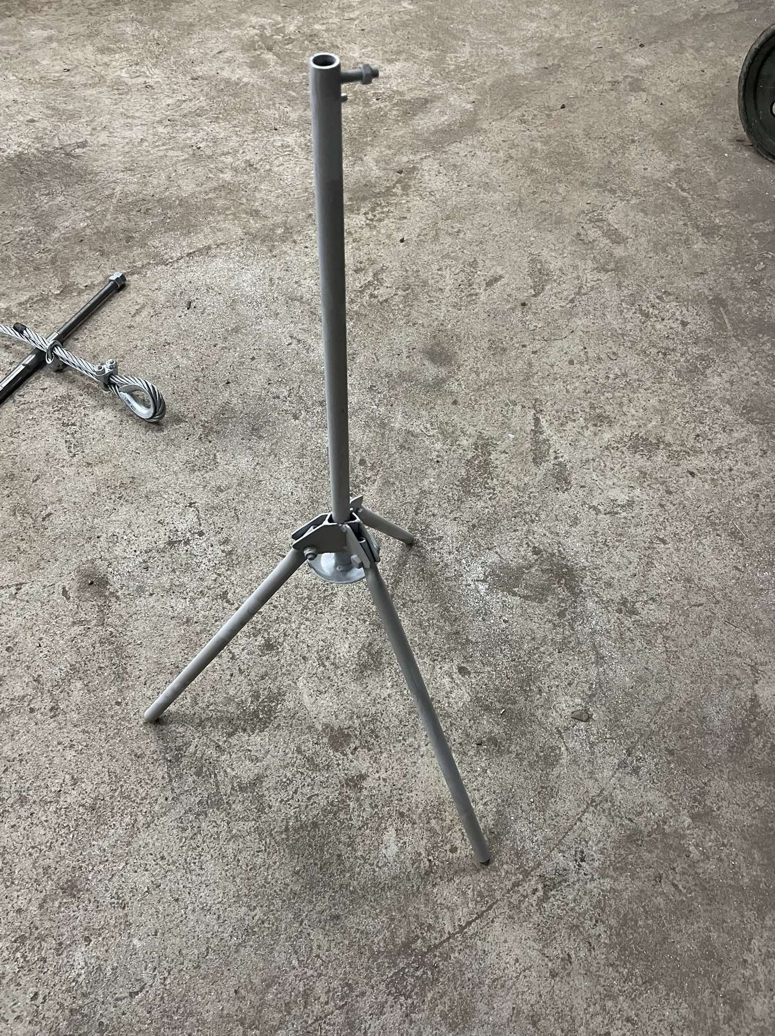 Szary, metalowy trójnóg z rurką na betonowym podłożu, widoczne elementy mocujące i lina stalowa w tle. Konstrukcja spawana, surowy wygląd.