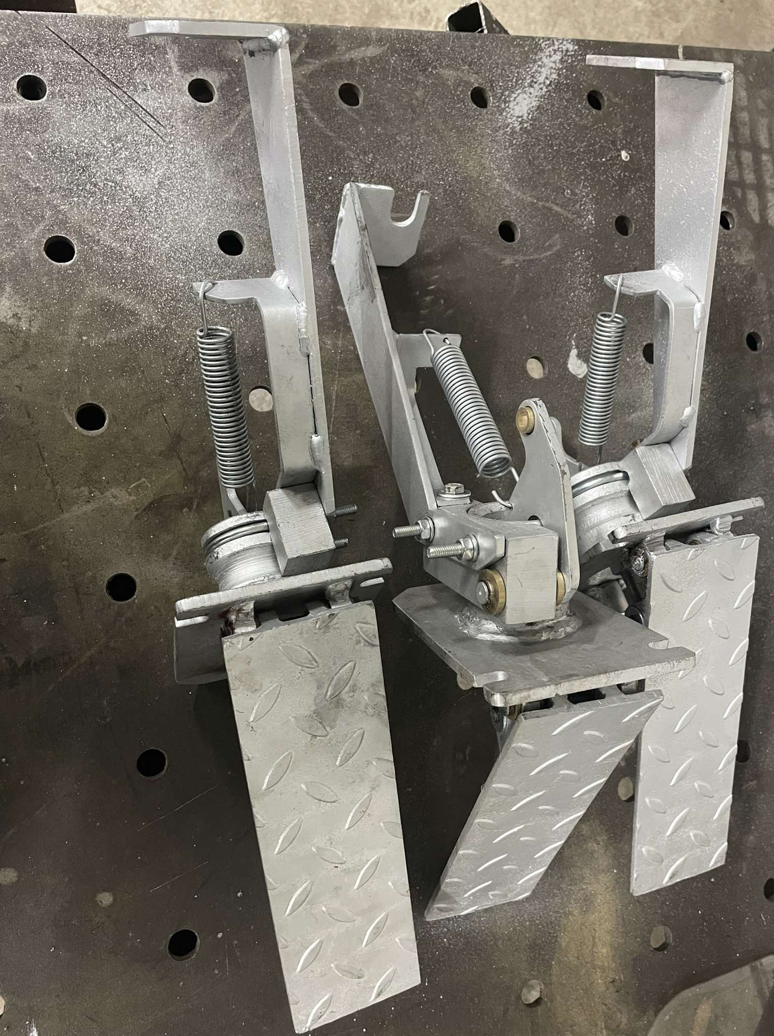 Dwa srebrne, metalowe pedały z sprężynami na perforowanym stole spawalniczym. Widoczne ślady obróbki i spawania. Detal konstrukcyjny z bliska.