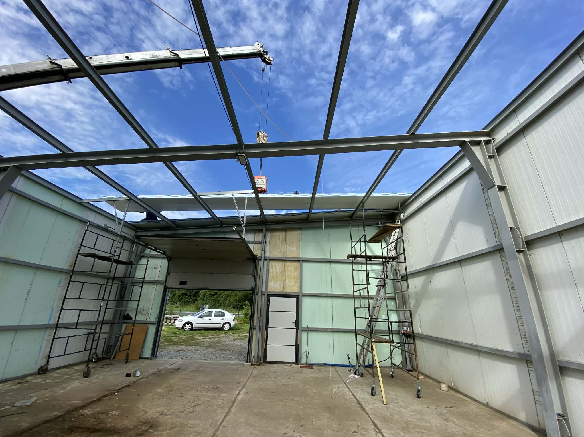 Wnętrze hali w budowie z widoczną stalową konstrukcją dachu i ścianami z płyt warstwowych. Na pierwszym planie rusztowania i podnośnik. W tle błękitne niebo.