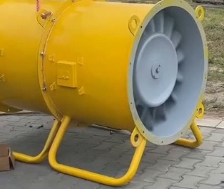 Żółty, przemysłowy wentylator osiowy na metalowej podstawie, z widocznymi łopatkami wirnika, zawieszony na haku dźwigu, gotowy do montażu w hali.