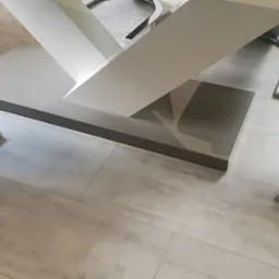 Szare płytki podłogowe ułożone w prostokątnym wzorze, widoczne fragmenty nowoczesnego stołu z białą podstawą i szarym blatem oraz krzesła z szarym siedziskiem i chromowanymi nogami.