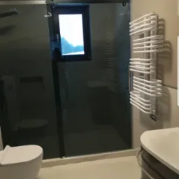 Nowoczesna łazienka z kabiną prysznicową typu walk-in z ciemną szybą, grzejnikiem drabinkowym i umywalką nablatową na szafce w odcieniach szarości.