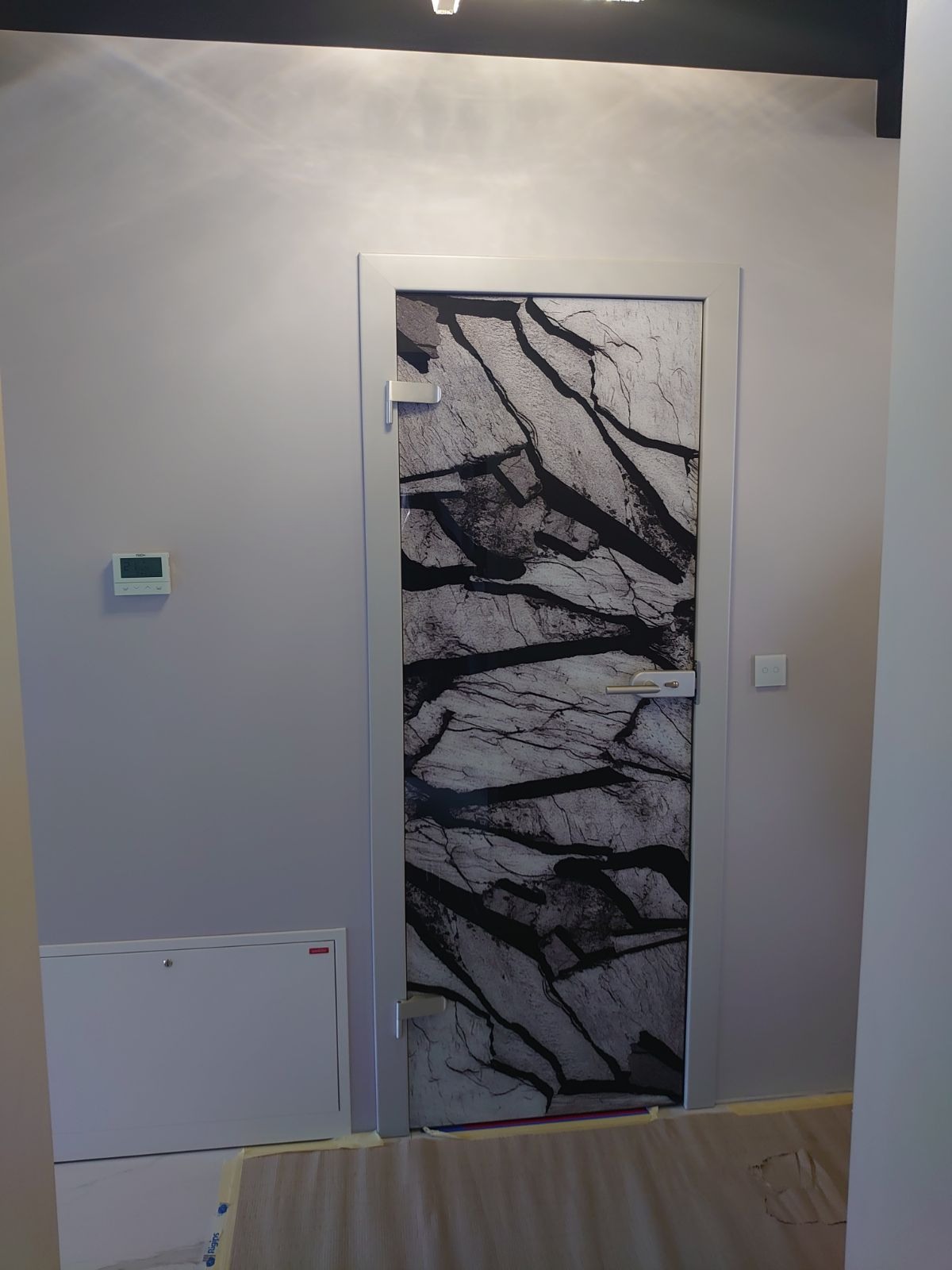 Drzwi wewnętrzne z okleiną imitującą marmur, w szarej ościeżnicy, zamontowane w szarym pomieszczeniu z widocznym termostatem i skrzynką podłączeniową, zabezpieczone folią malarską na podłodze.