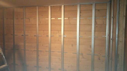 Konstrukcja metalowa mocowana do drewnianej ściany z desek w trakcie remontu wnętrza, widoczne profile pionowe i poziome mocowania.