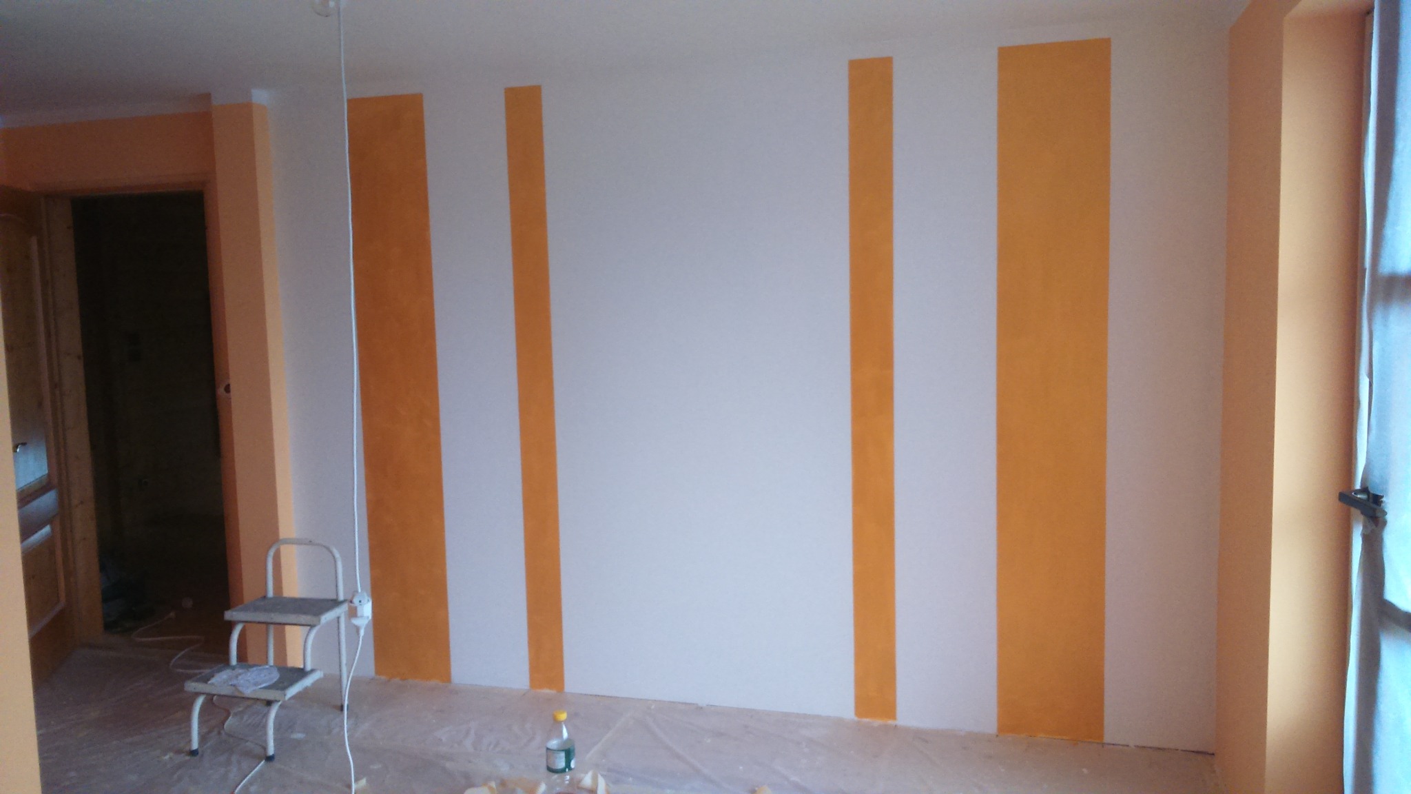 Ściana w pokoju z pomalowanymi pionowymi pasami w kolorze pomarańczowym i białym; na podłodze folia ochronna i drabinka malarska.