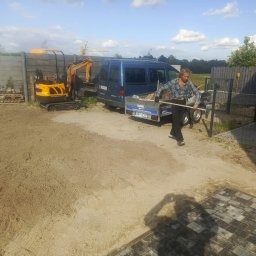 SAVA - Pracownik z grabiami na placu budowy. W tle mini koparka, przyczepa i ogrodzenie betonowe. Częściowo ułożona kostka brukowa. Słoneczny dzień.