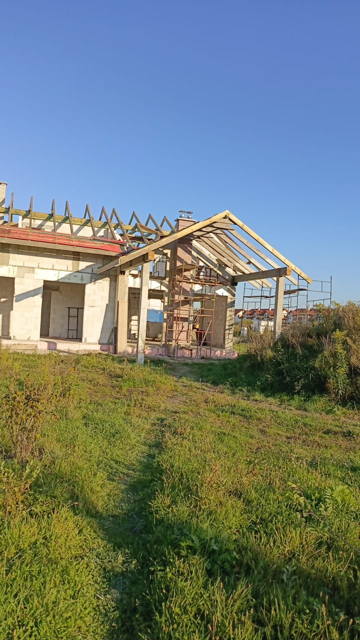 Surowy stan domu w trakcie budowy z widoczną konstrukcją dachu, murowanymi ścianami i rusztowaniami na zewnątrz, otoczony zielenią i błękitnym niebem.