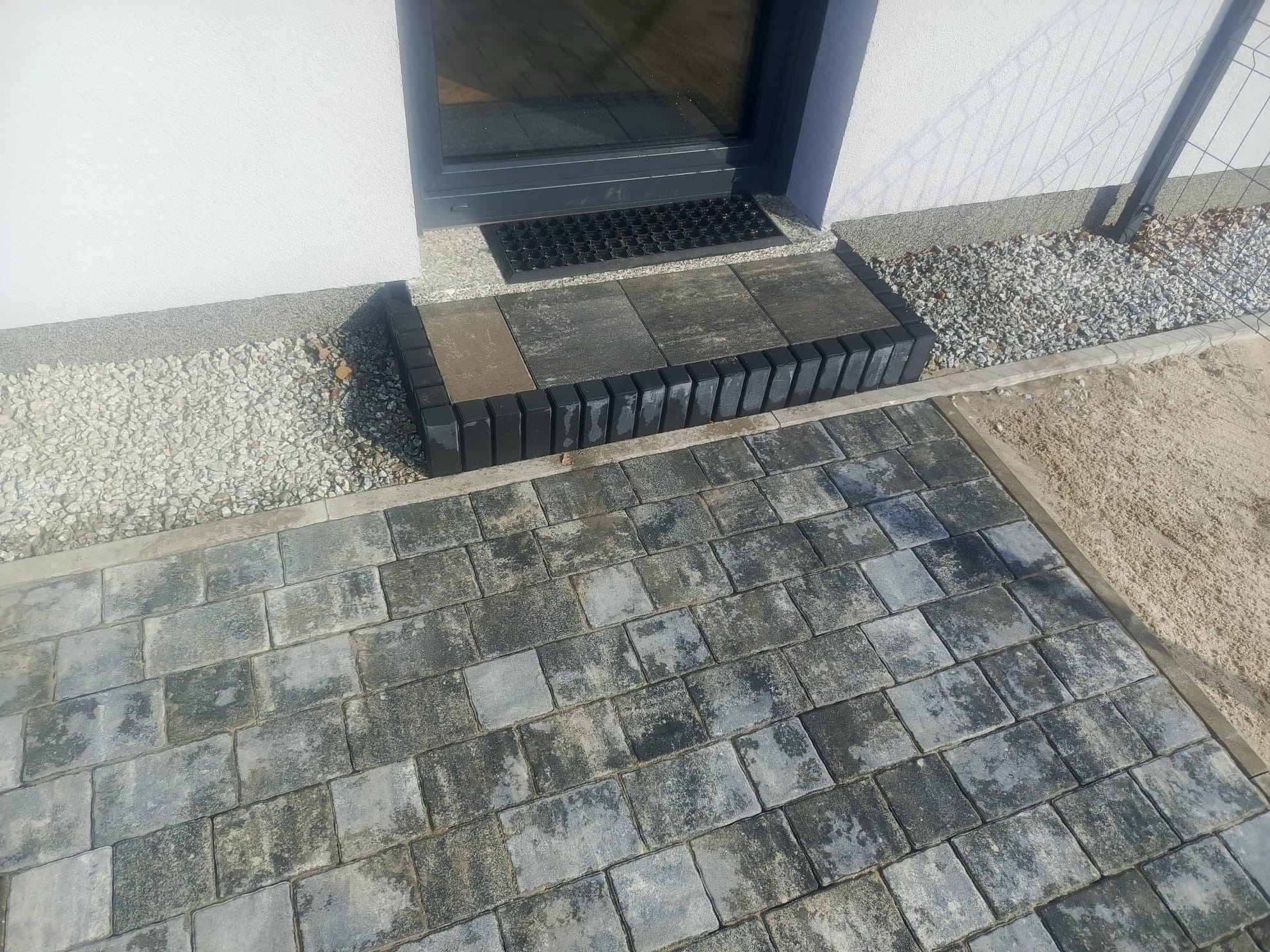 Ułożona kostka brukowa w Kamieńcu Wrocławskim, tworząca ścieżkę i schody wejściowe do budynku. Widoczne szare kostki i czarne obrzeża. Wykończenie z kamyków i piasku.