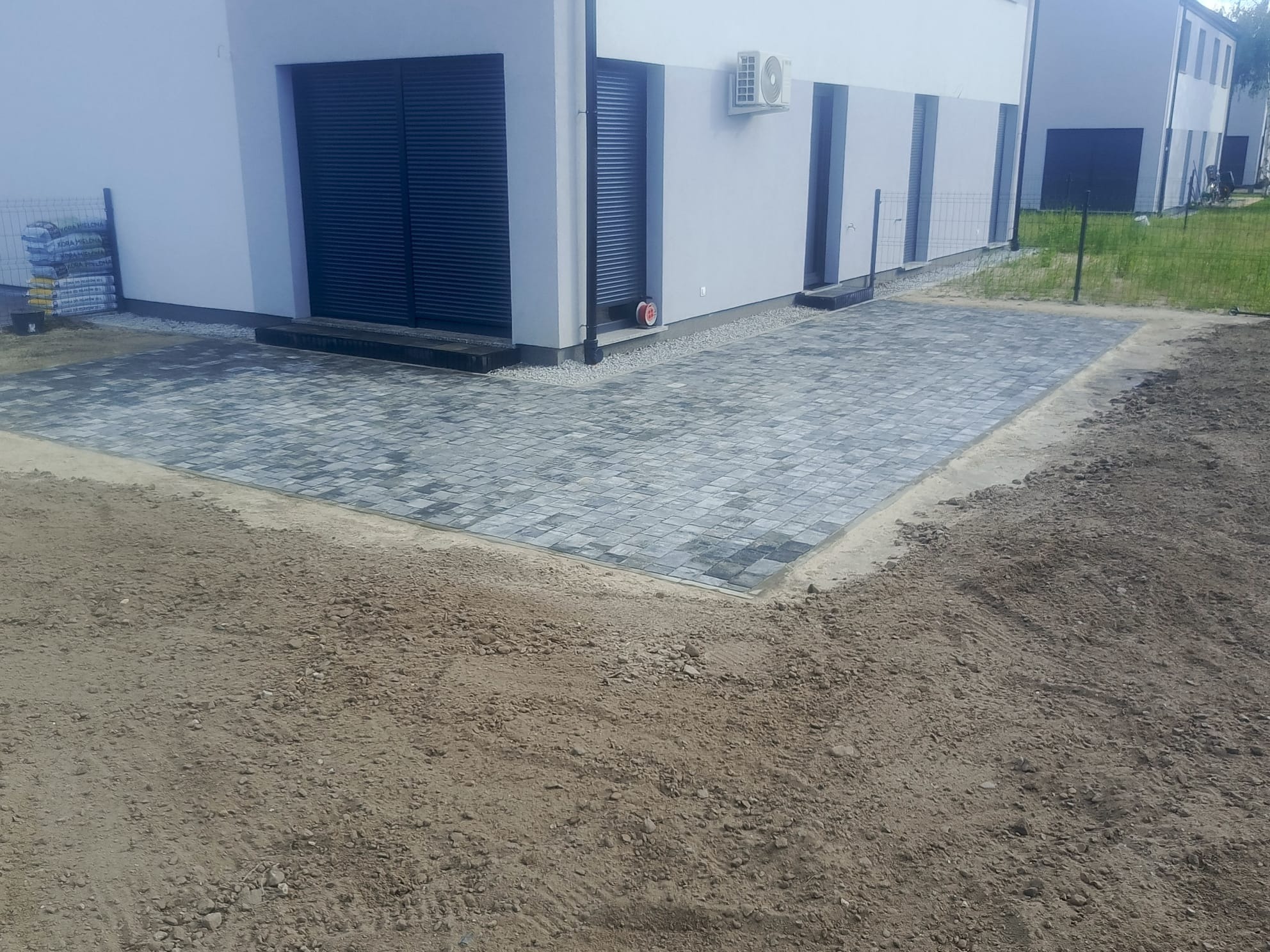 Szara kostka brukowa ułożona przed nowoczesnym domem z roletami. Częściowo przygotowany teren pod dalsze prace, widoczne worki z materiałami budowlanymi.