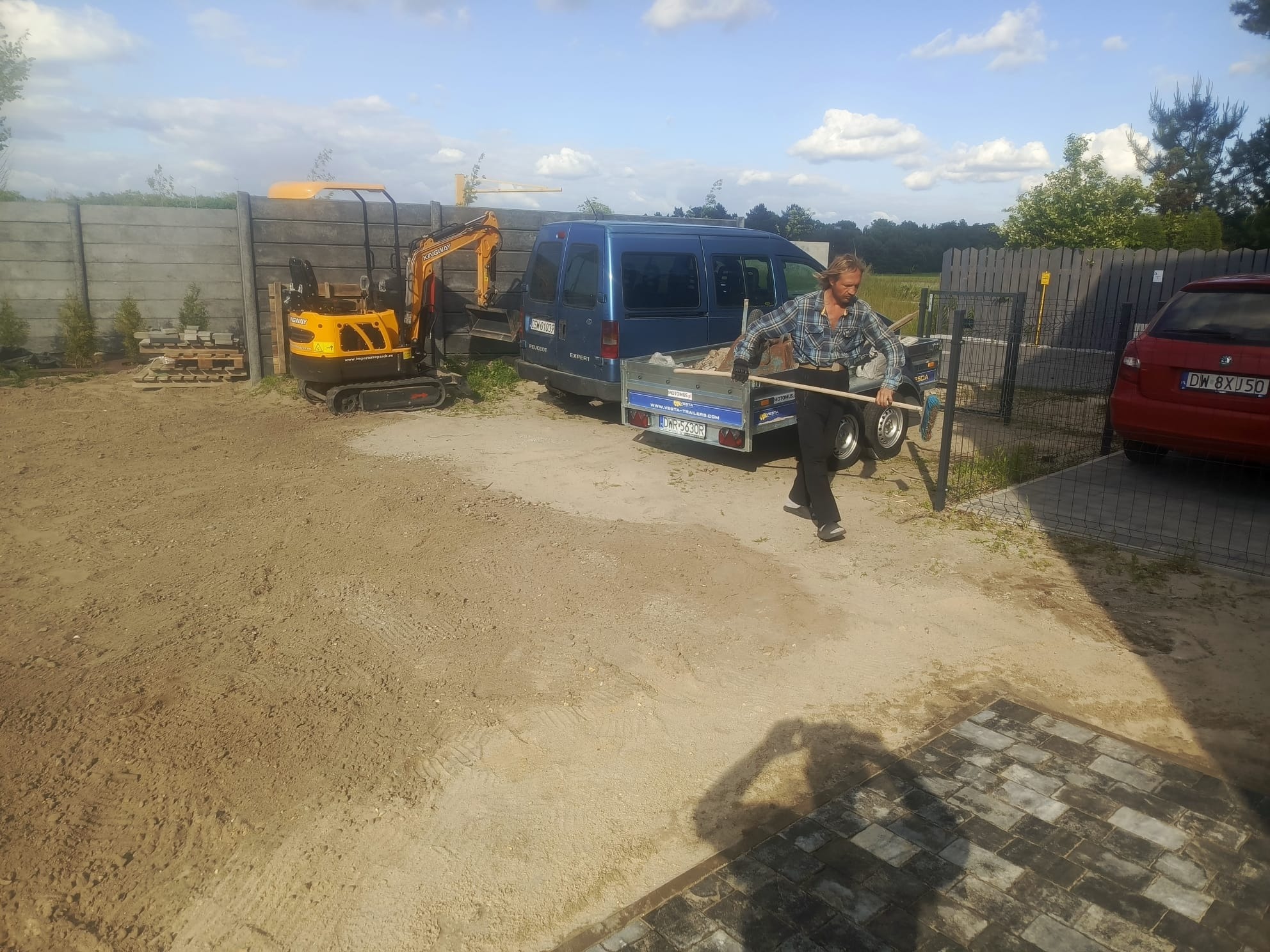 Pracownik z grabiami na placu budowy. W tle mini koparka, przyczepa i ogrodzenie betonowe. Częściowo ułożona kostka brukowa. Słoneczny dzień.
