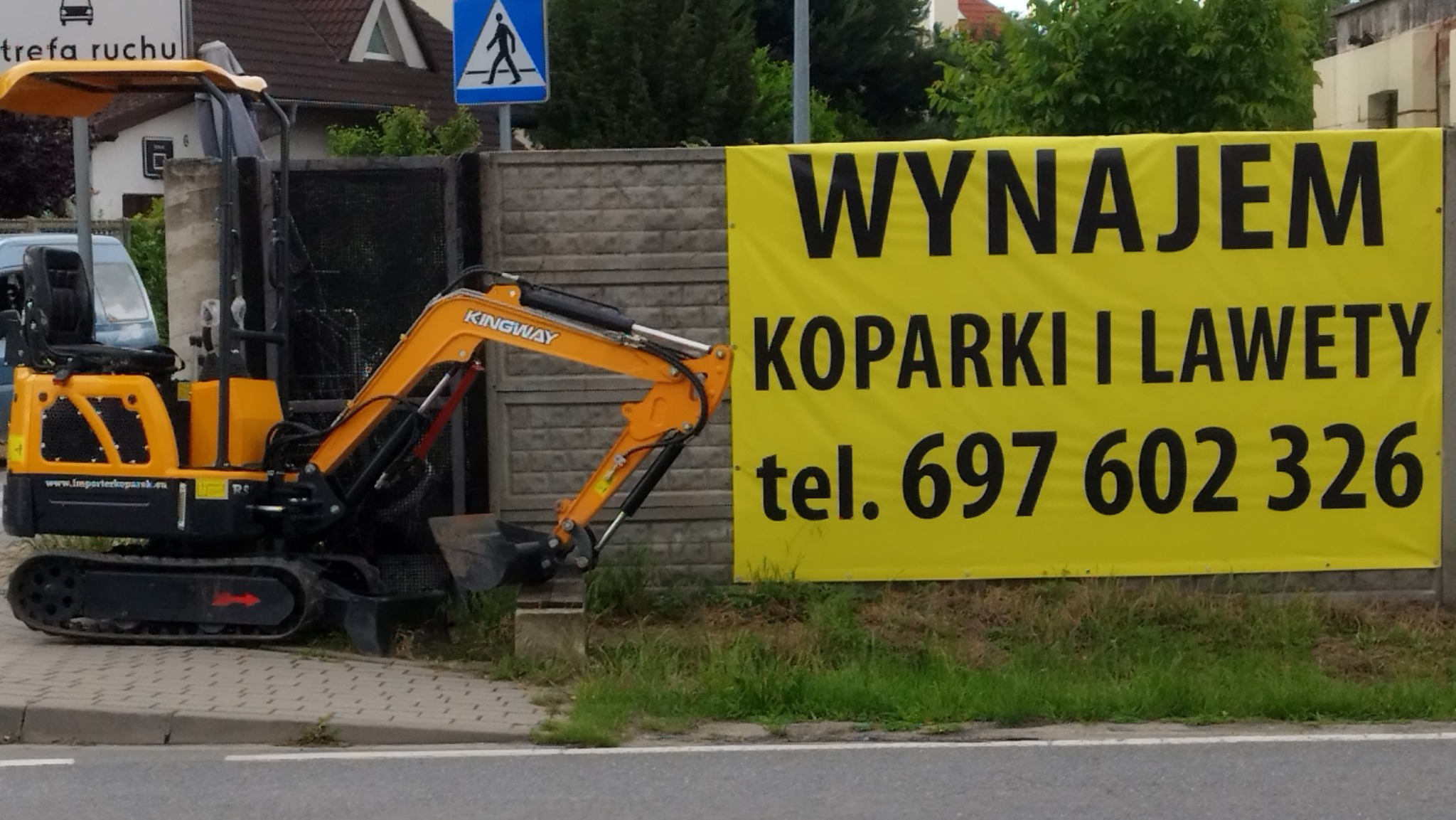 Żółta mini koparka Kingway na chodniku obok baneru z napisem 'Wynajem koparki i lawety' oraz numerem telefonu, widok z boku.