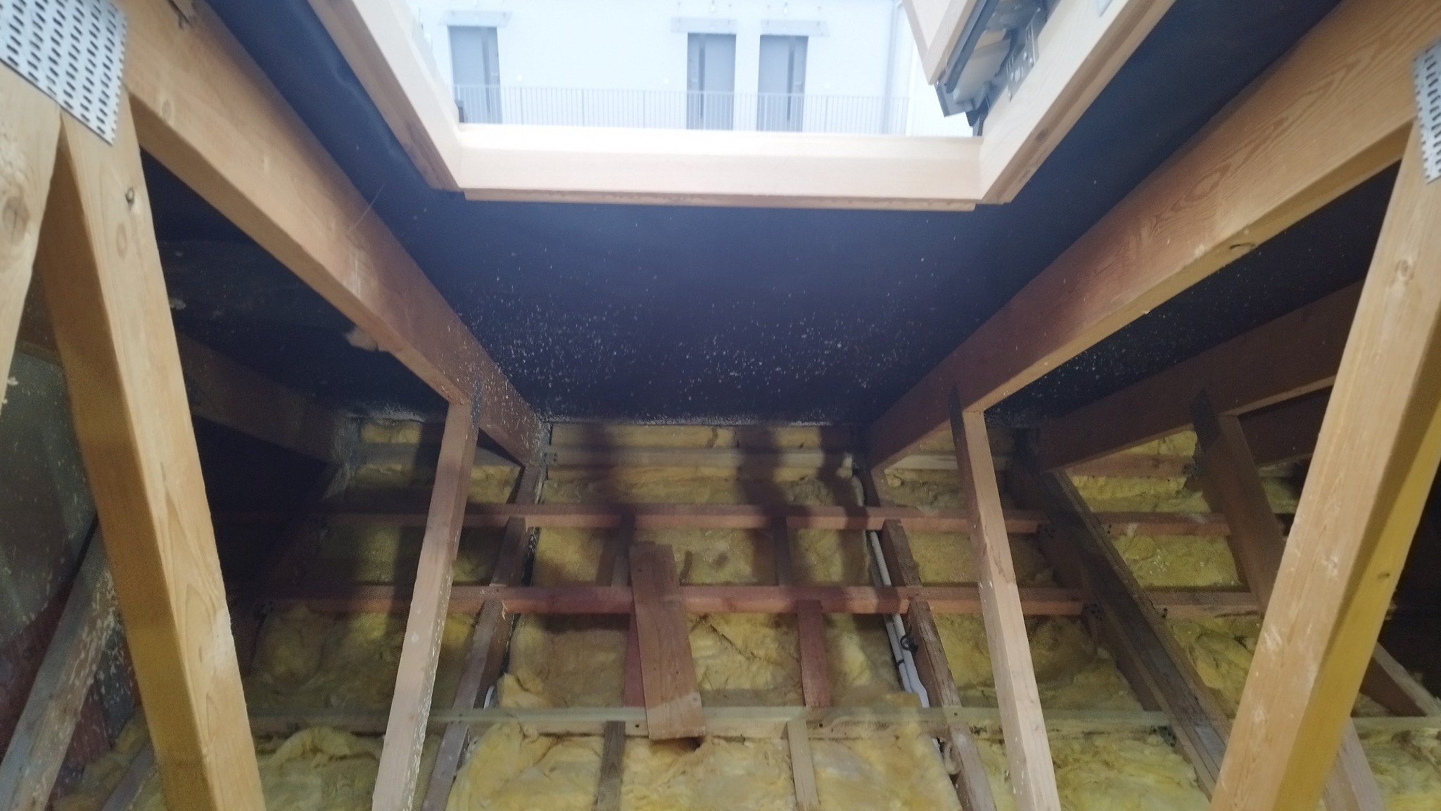 Skośny dach z oknem połaciowym w trakcie ocieplania. Widoczne drewniane belki konstrukcyjne i żółta wełna mineralna. Na zewnątrz widok na budynek z oknami.