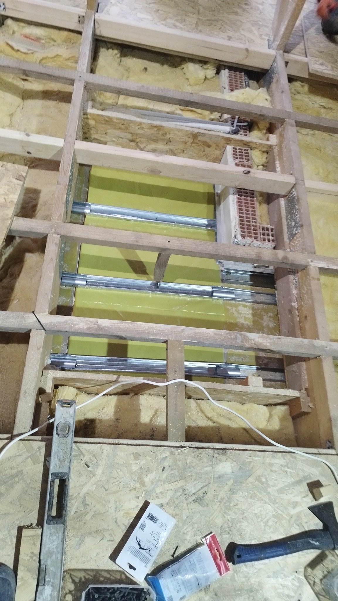 Szkielet drewniany z izolacją, widoczne profile metalowe i fragment muru. Poziomica i narzędzia na podłodze. Ujęcie z góry, ukazujące etap prac budowlanych.