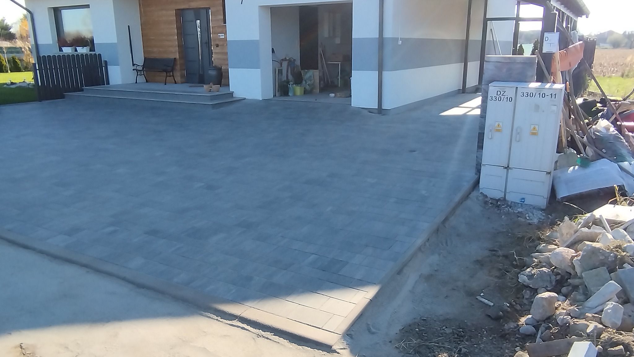 Ułożona szara kostka brukowa na podjeździe przed nowoczesnym domem z garażem. Widoczne obrzeża i skrzynka elektryczna. Częściowo widoczne gruz i materiały budowlane.