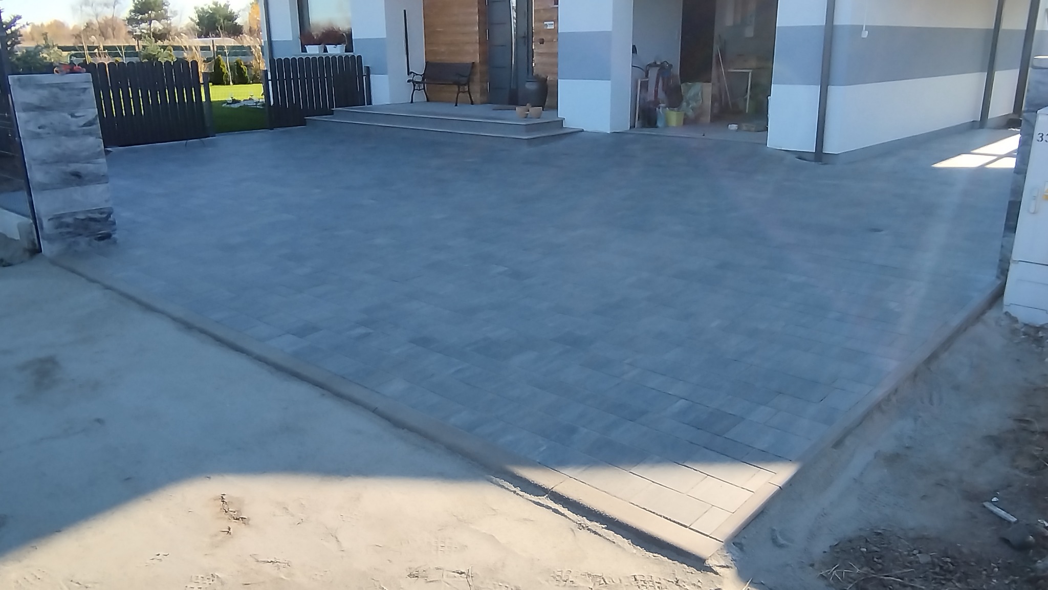 Szara kostka brukowa ułożona na podjeździe przed domem z elementami ogrodzenia i schodami wejściowymi. Widok z góry, lekko z boku, prezentujący wzór ułożenia.