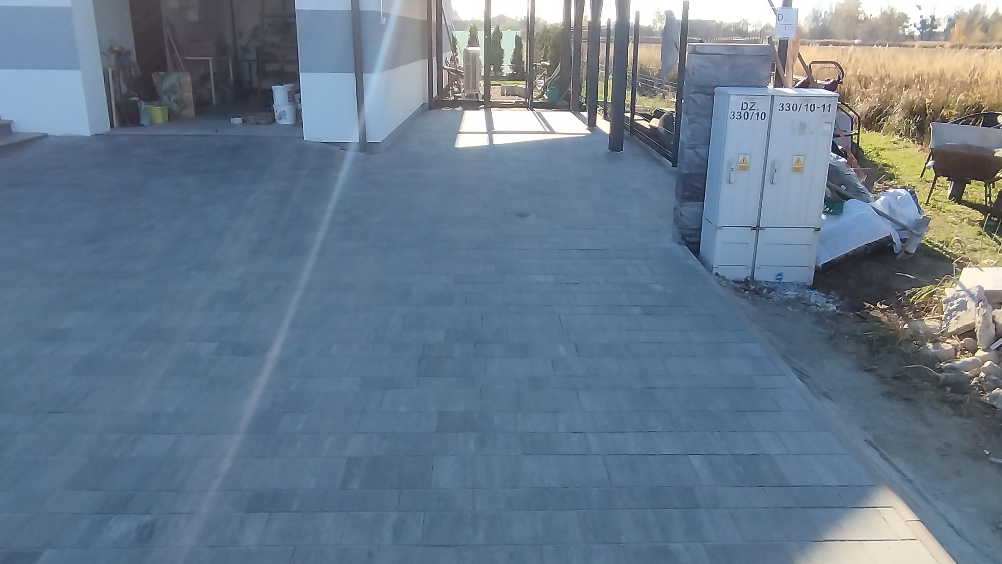 Ułożona szara kostka brukowa na podjeździe, z widoczną skrzynką elektryczną i elementami ogrodu w tle, w słoneczny dzień. Widok z perspektywy.