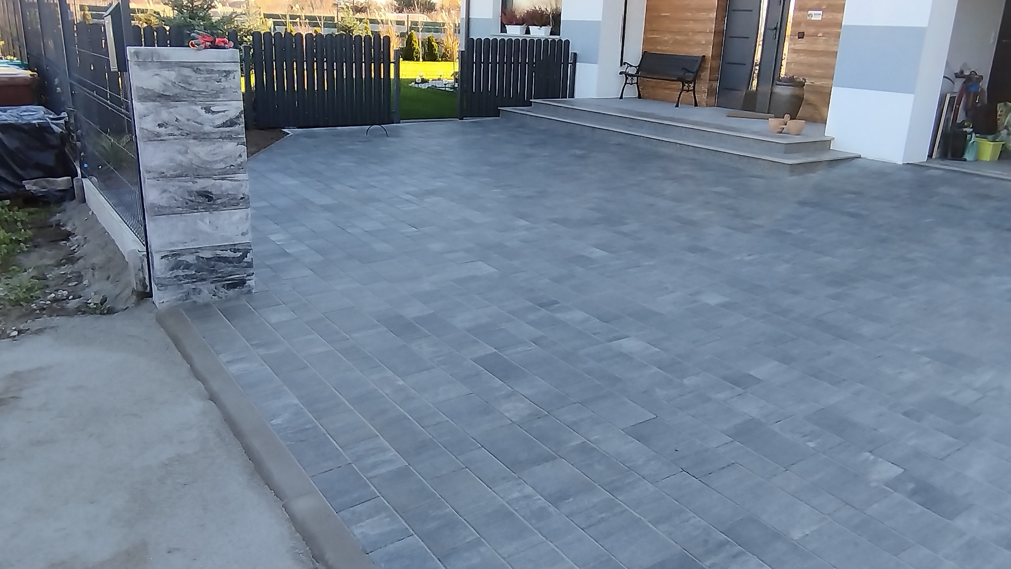 Ułożona szara kostka brukowa na podjeździe, widok na bramę i wejście do domu. Estetyczne wykończenie, równa powierzchnia, schludny wygląd.