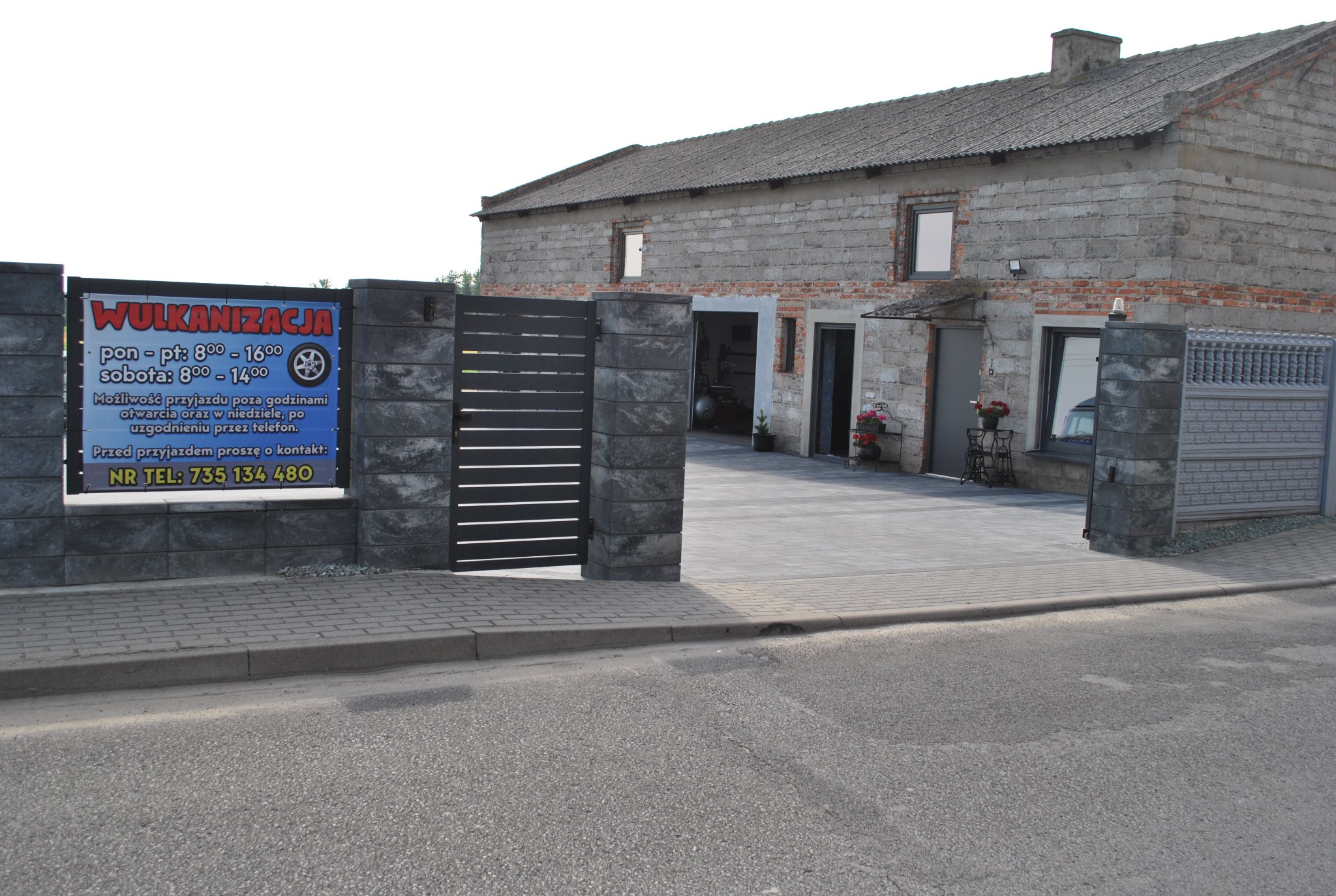Budynek wulkanizacji z szarej cegły, brama wjazdowa z szarych bloków i tablicą informacyjną o godzinach otwarcia i numerze telefonu.