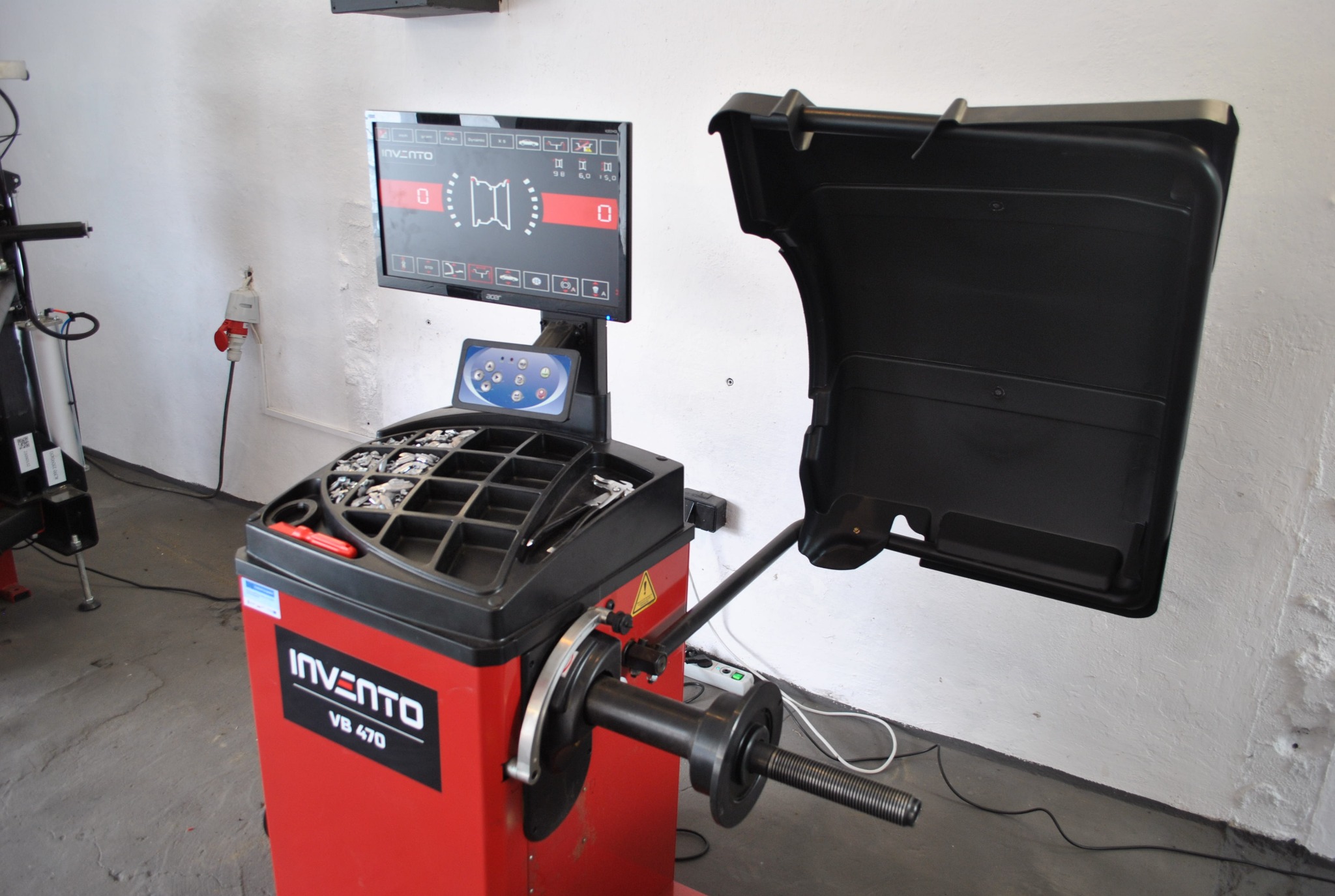 Wyważarka do kół Invento VB 470 z ekranem i osłoną, widoczna w warsztacie samochodowym, gotowa do użycia. Na ekranie widoczne parametry ustawień.