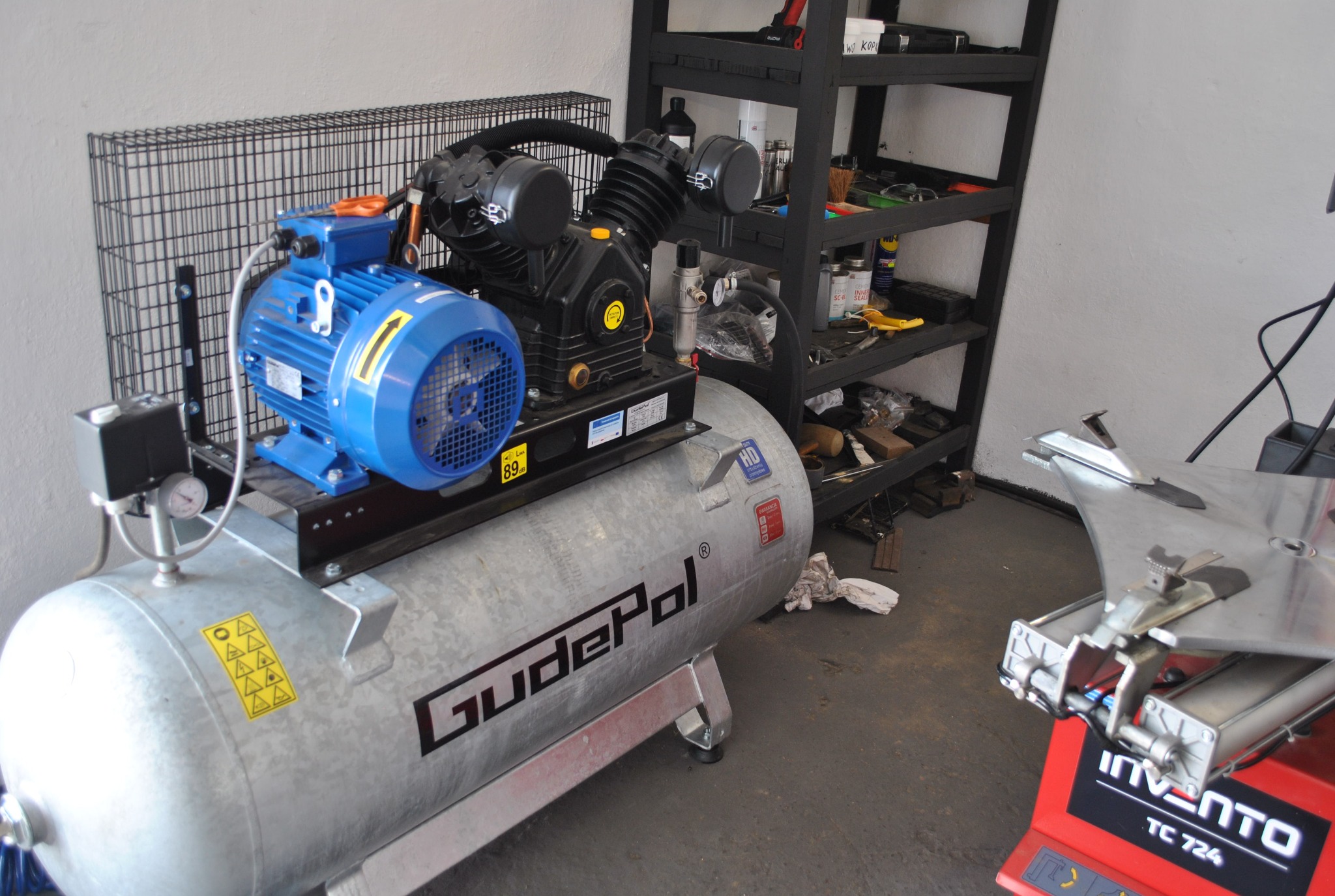 Kompresor GudePol z niebieskim silnikiem, stojący w warsztacie obok wyważarki do kół Invento TC 724, z półkami na narzędzia w tle.