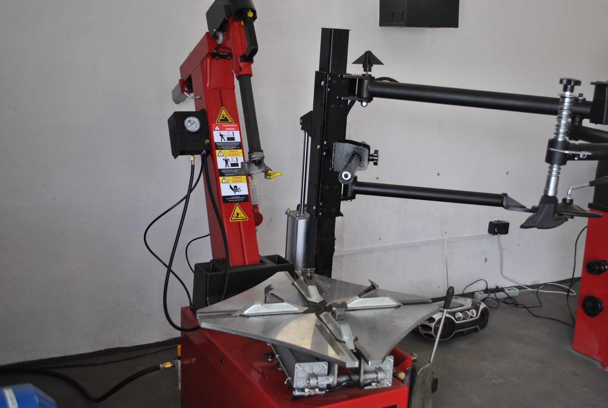 Czerwono-czarna montażownica do opon z manometrem i ostrzeżeniami BHP na tle szarej ściany warsztatu, obok widoczny radioodtwarzacz i plątanina kabli.