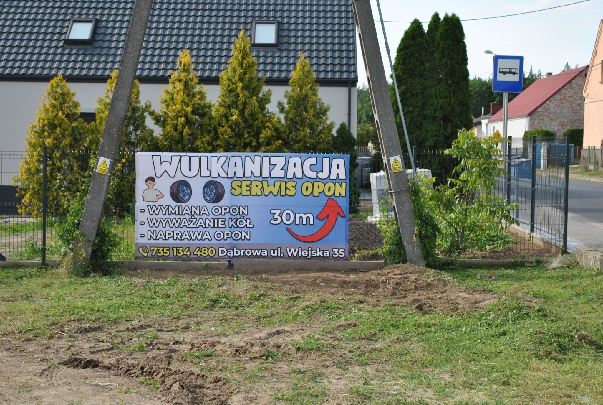 Baner reklamowy wulkanizacji i serwisu opon, umieszczony na tle zieleni i słupów betonowych, z widoczną informacją o wymianie, wyważaniu i naprawie opon oraz odległości 30m.