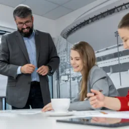 Mężczyzna w garniturze prezentuje diagram na flipcharcie grupie osób siedzących przy stole podczas szkolenia biznesowego; widoczna filiżanka kawy, tablet i notatki.