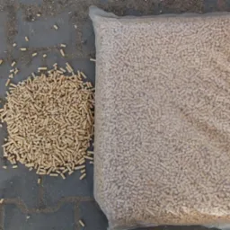 Worek przezroczysty wypełniony pelletem drzewnym leży obok rozsypanej kupki pelletu na szarej kostce brukowej.