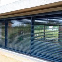 Zamknięte, grafitowe żaluzje zewnętrzne zamontowane w oknie budynku w trakcie budowy, widoczne elementy konstrukcyjne i piasek.