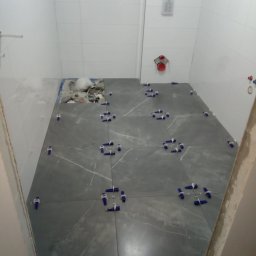 ROBI-HOME Posadzki Żywice Kamienny Dywan - Szare płytki podłogowe z białym wzorem żyłkowania, ułożone w łazience, z użyciem niebieskich klinów do regulacji odstępów, widoczne białe płytki ścienne i instalacja wodna w tle.