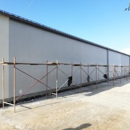 ROBI-HOME Posadzki Żywice Kamienny Dywan - Długa ściana budynku przemysłowego po nałożeniu szarej warstwy wykończeniowej, widoczne rusztowania wzdłuż ściany, błękitne niebo w tle.