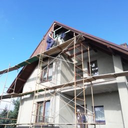 ROBI-HOME Posadzki Żywice Kamienny Dywan - Pracownik na rusztowaniu montuje elementy wykończeniowe pod brązowym dachem domu jednorodzinnego, widok z dołu na tle błękitnego nieba.