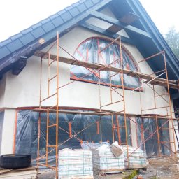 ROBI-HOME Posadzki Żywice Kamienny Dywan - Dom jednorodzinny w trakcie prac wykończeniowych elewacji: nałożona warstwa szpachli, okna zabezpieczone folią, rozstawione rusztowania, w tle widoczny las.