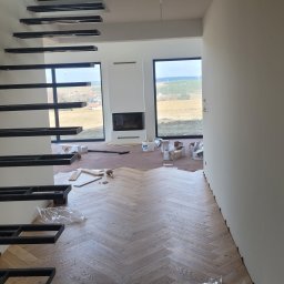 ROBI-HOME Posadzki Żywice Kamienny Dywan - Wnętrze w trakcie wykańczania: ułożona podłoga z paneli w jodełkę, surowe schody wspornikowe i przygotowane ściany w nowoczesnym domu z dużymi oknami.