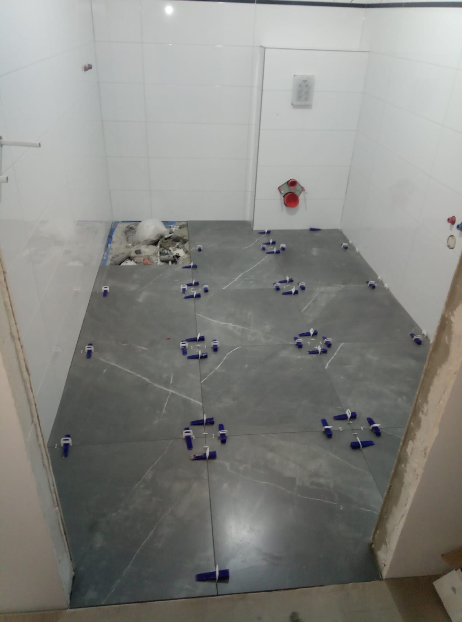 Szare płytki podłogowe z białym wzorem żyłkowania, ułożone w łazience, z użyciem niebieskich klinów do regulacji odstępów, widoczne białe płytki ścienne i instalacja wodna w tle.