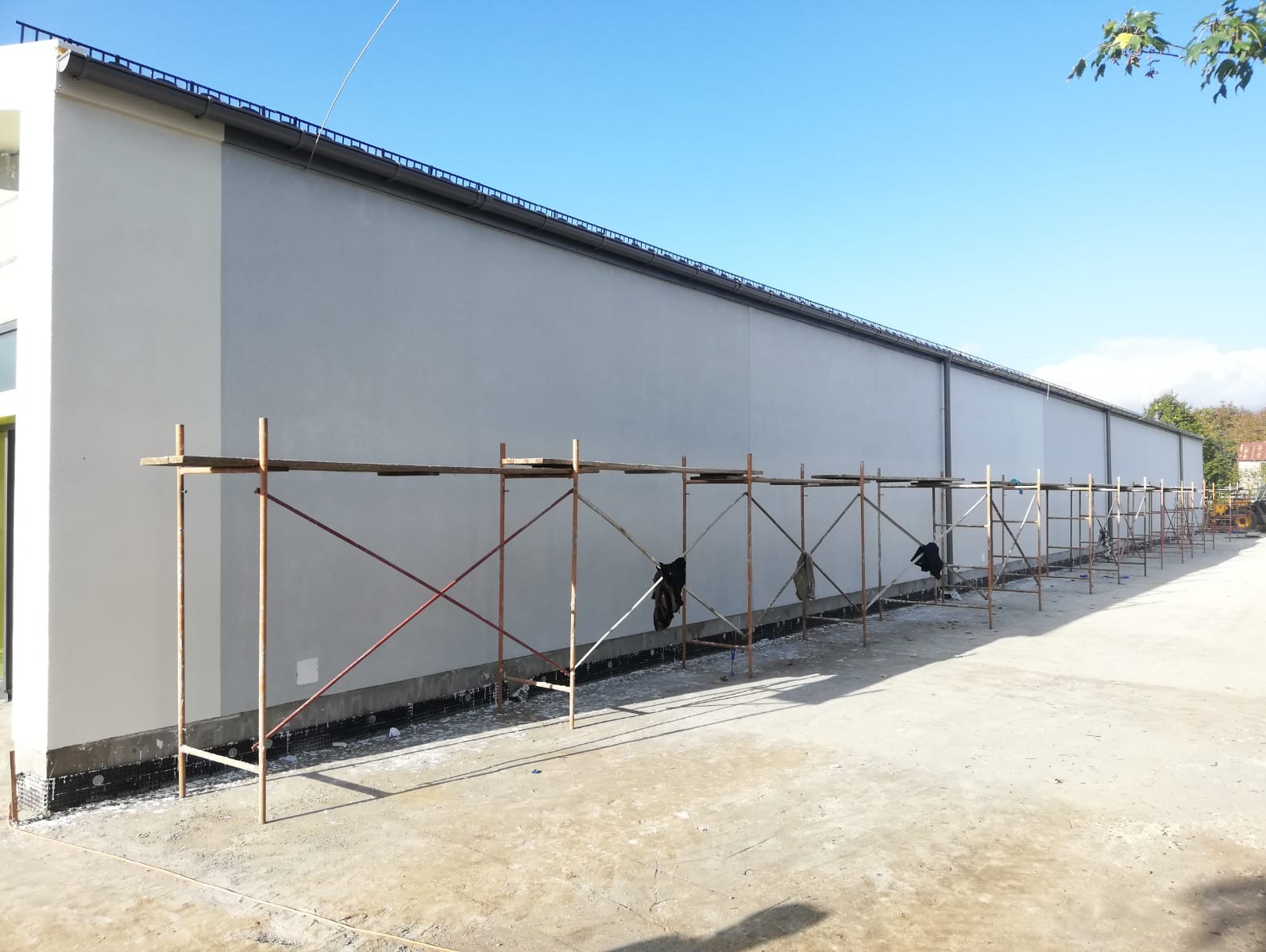 Długa ściana budynku przemysłowego po nałożeniu szarej warstwy wykończeniowej, widoczne rusztowania wzdłuż ściany, błękitne niebo w tle.