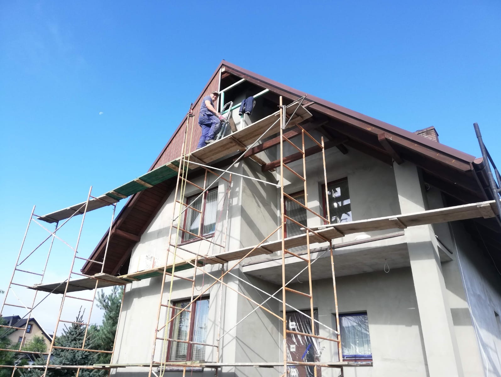 Pracownik na rusztowaniu montuje elementy wykończeniowe pod brązowym dachem domu jednorodzinnego, widok z dołu na tle błękitnego nieba.