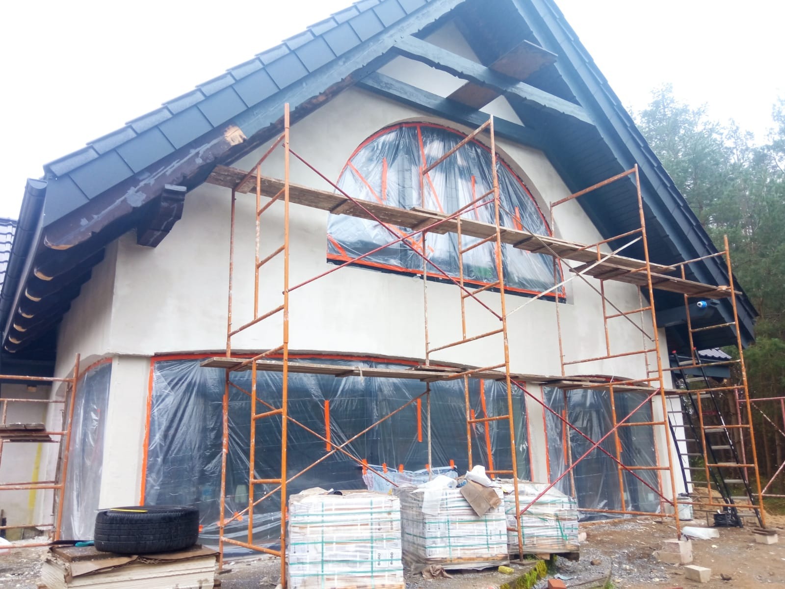 Dom jednorodzinny w trakcie prac wykończeniowych elewacji: nałożona warstwa szpachli, okna zabezpieczone folią, rozstawione rusztowania, w tle widoczny las.