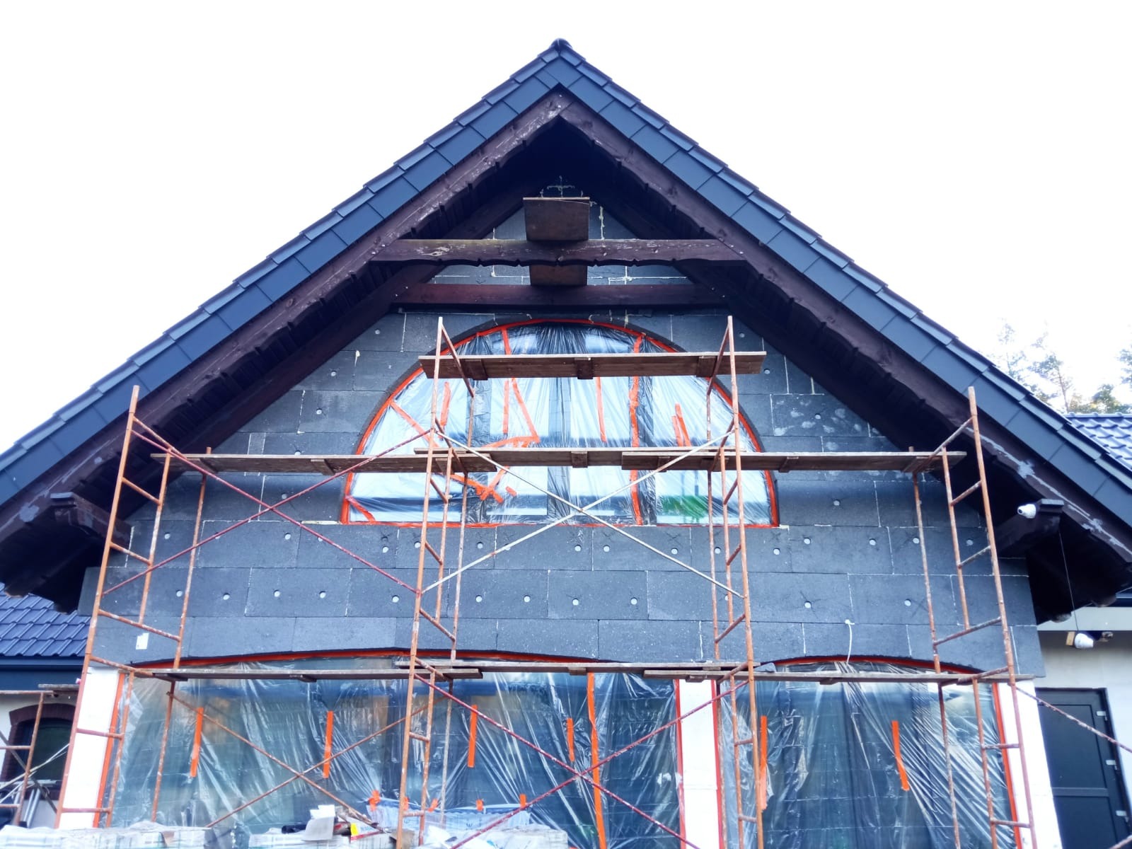 Budynek w trakcie ocieplania styropianem grafitowym, zabezpieczony folią i rusztowaniem, widoczne drewniane elementy konstrukcyjne dachu.