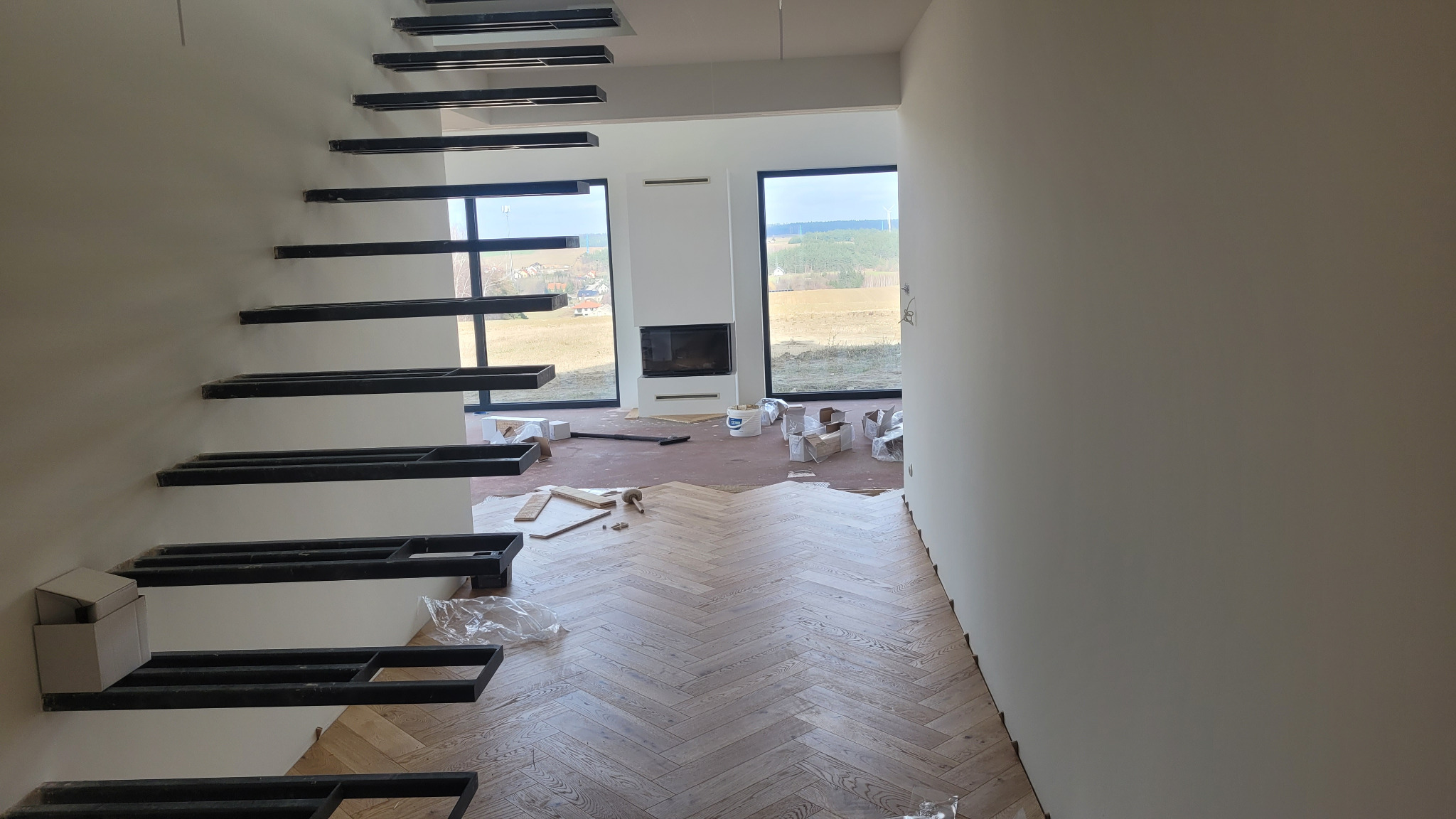 Wnętrze w trakcie wykańczania: ułożona podłoga z paneli w jodełkę, surowe schody wspornikowe i przygotowane ściany w nowoczesnym domu z dużymi oknami.