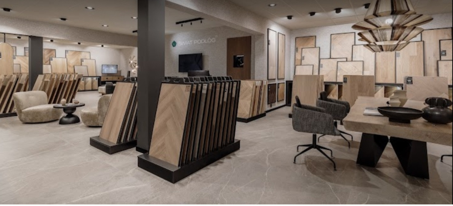 Ekspozycja salonu z różnymi wzorami paneli podłogowych ułożonymi w stojakach, widoczne także próbki na ścianach, meble wypoczynkowe i stół z dekoracjami, całość w stonowanej kolorystyce.