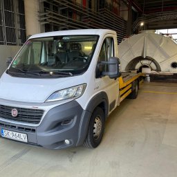 Auto Handel BAJCAR - Biały bus z żółtą lawetą przewozi duży, metalowy element konstrukcyjny w hali przemysłowej z widocznymi rurami i instalacjami pod sufitem.