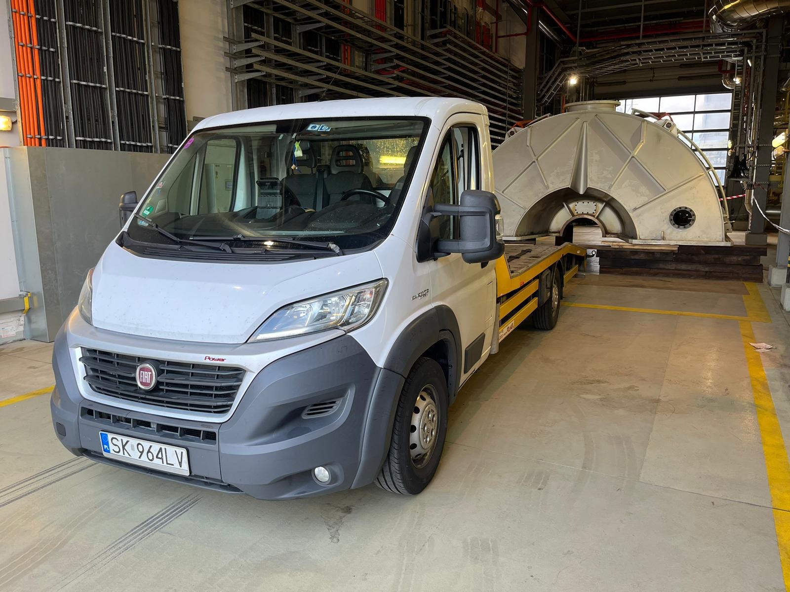 Biały bus z żółtą lawetą przewozi duży, metalowy element konstrukcyjny w hali przemysłowej z widocznymi rurami i instalacjami pod sufitem.