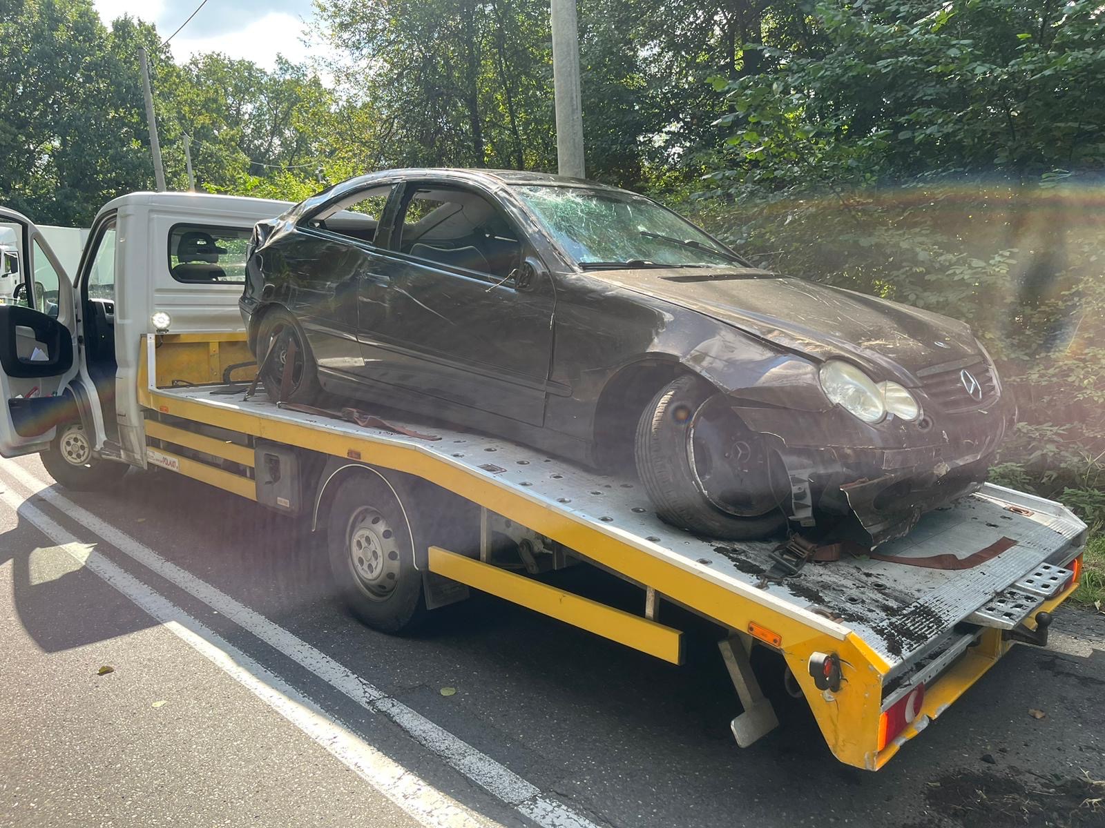 Uszkodzony, czarny Mercedes CLK na żółtej lawecie, z rozbitą przednią szybą i zniszczonym przodem, w tle zieleń drzew.