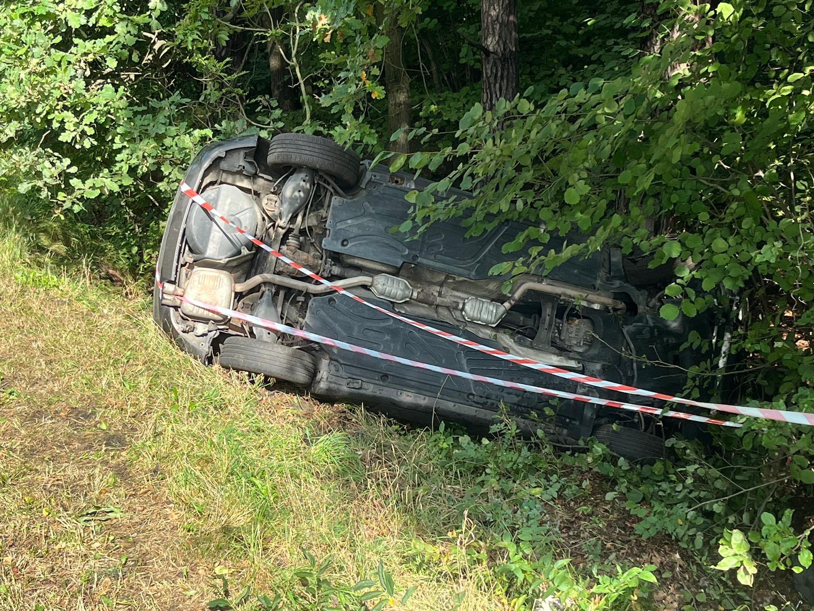 Przewrócony samochód na dachu w zaroślach, otoczony taśmą ostrzegawczą.