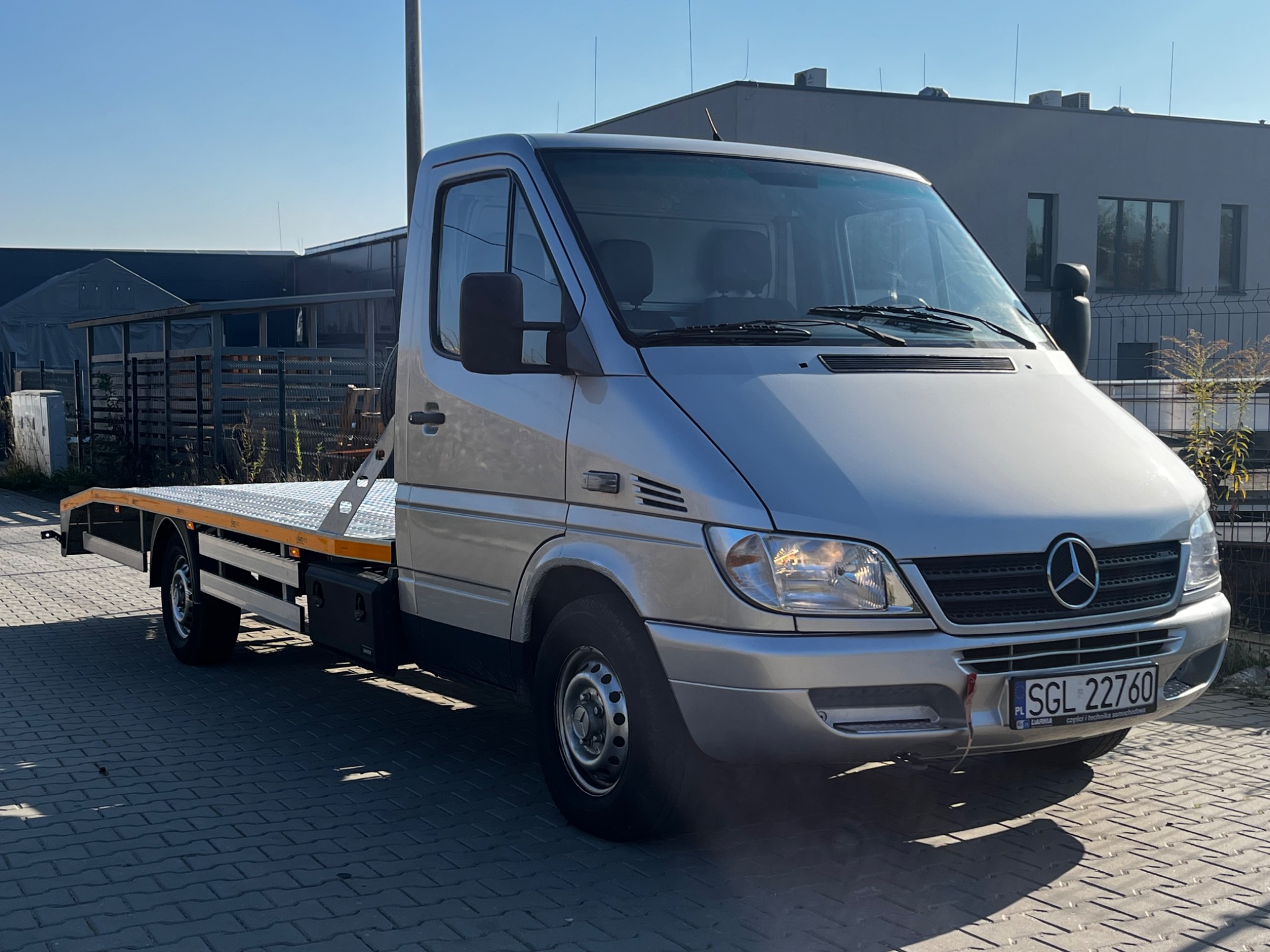 Srebrna laweta Mercedes-Benz z pustą platformą załadunkową, zaparkowana na brukowanej nawierzchni, z budynkiem w tle w słoneczny dzień.