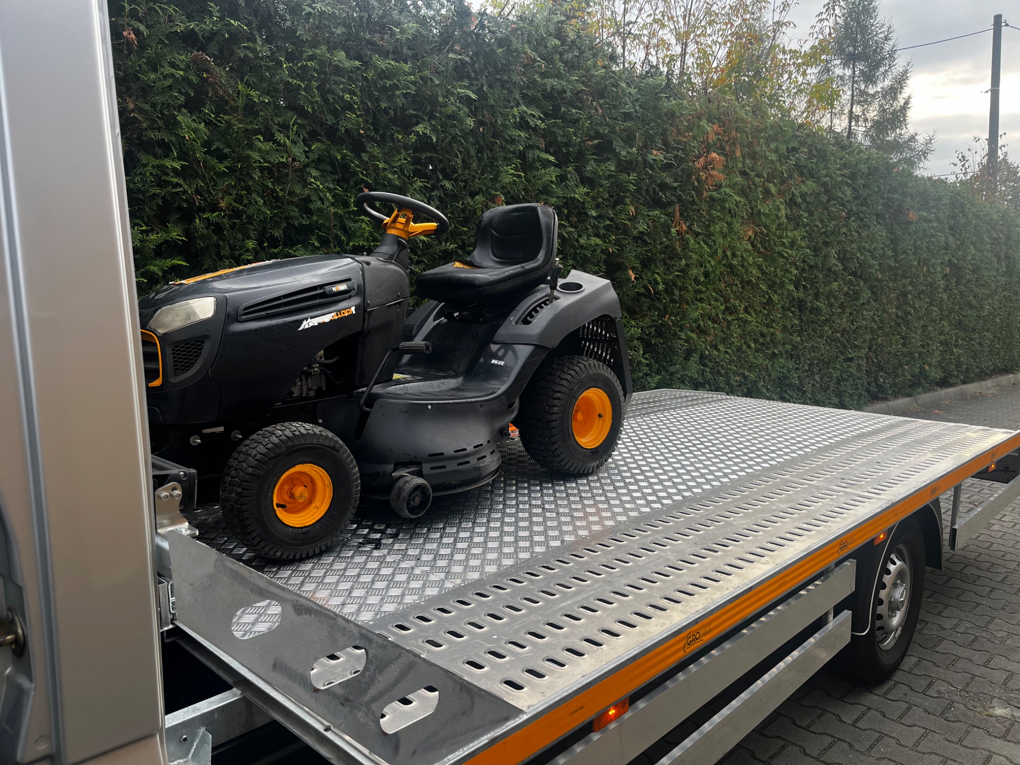 Czarny traktorek do koszenia trawy z żółtymi elementami, przewożony na srebrnej platformie lawety na tle zielonego żywopłotu.