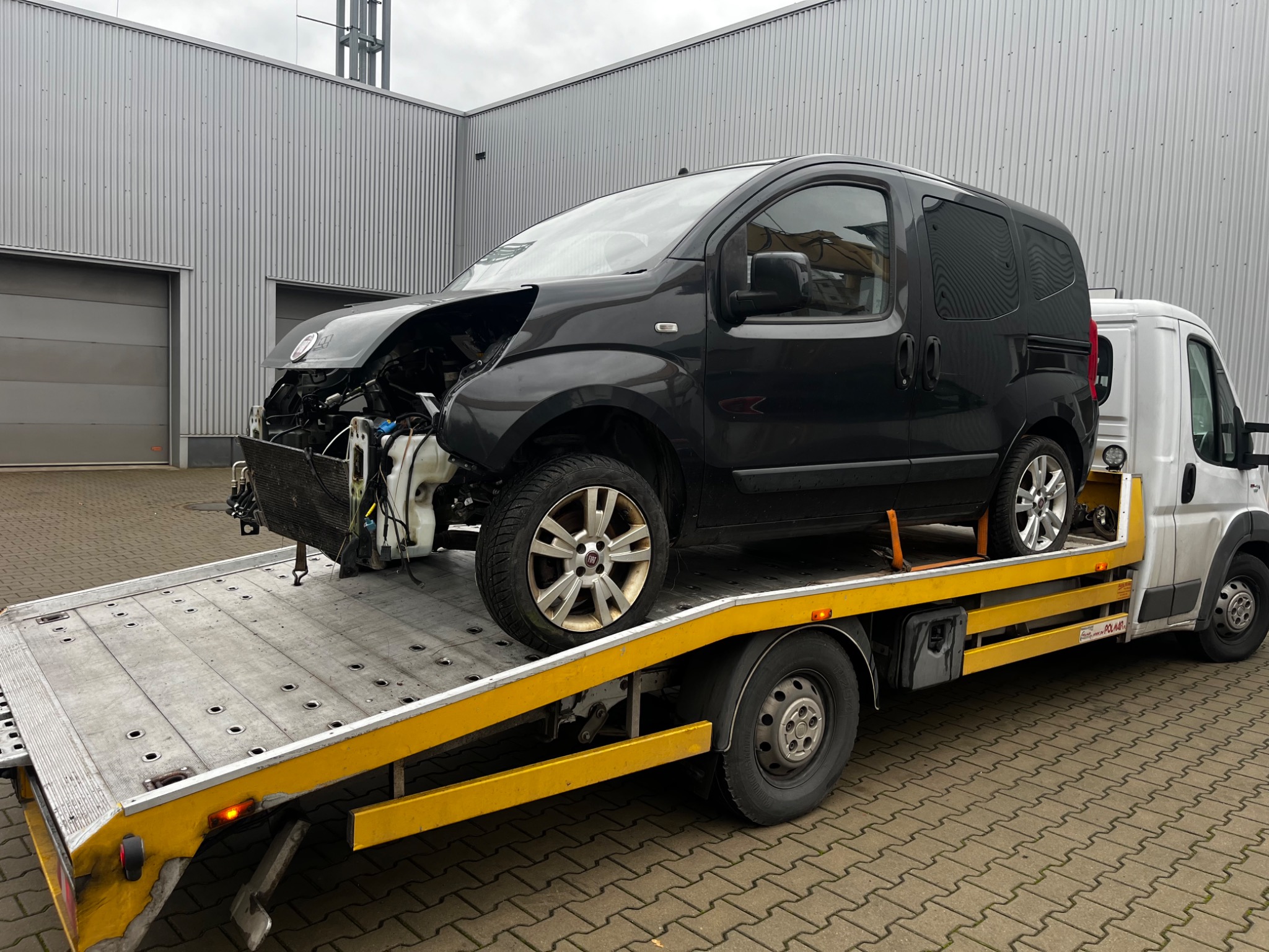 Uszkodzony, czarny bus na żółtej lawecie, widoczne braki w karoserii przodu, tło stanowi szary budynek przemysłowy.